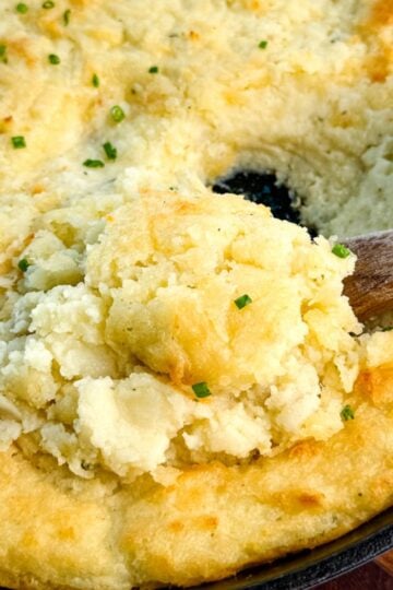 Close-up of a cast-iron skillet filled with golden, fluffy baked mashed potatoes topped with chives, with a wooden spoon scooping out a serving.