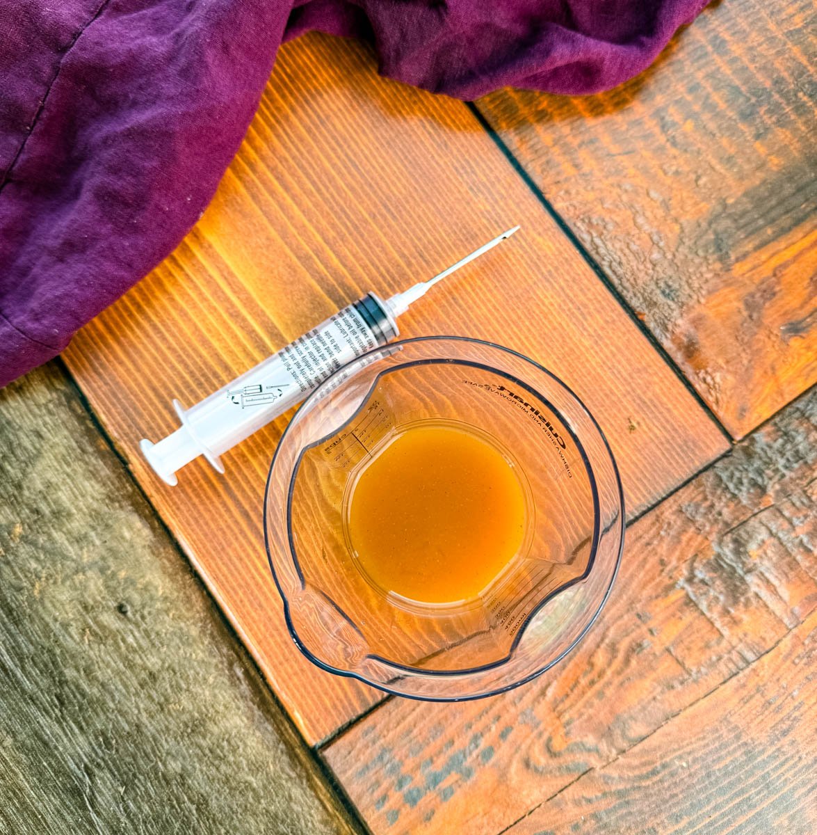 Measuring cup with seasoned injectable marinade and a marinade syringe on a wooden surface with a purple cloth.