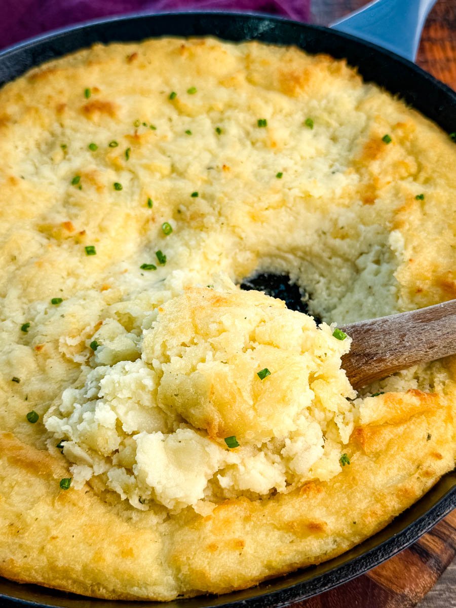 Close-up of a cast-iron skillet filled with golden, fluffy baked mashed potatoes topped with chives, with a wooden spoon scooping out a serving.