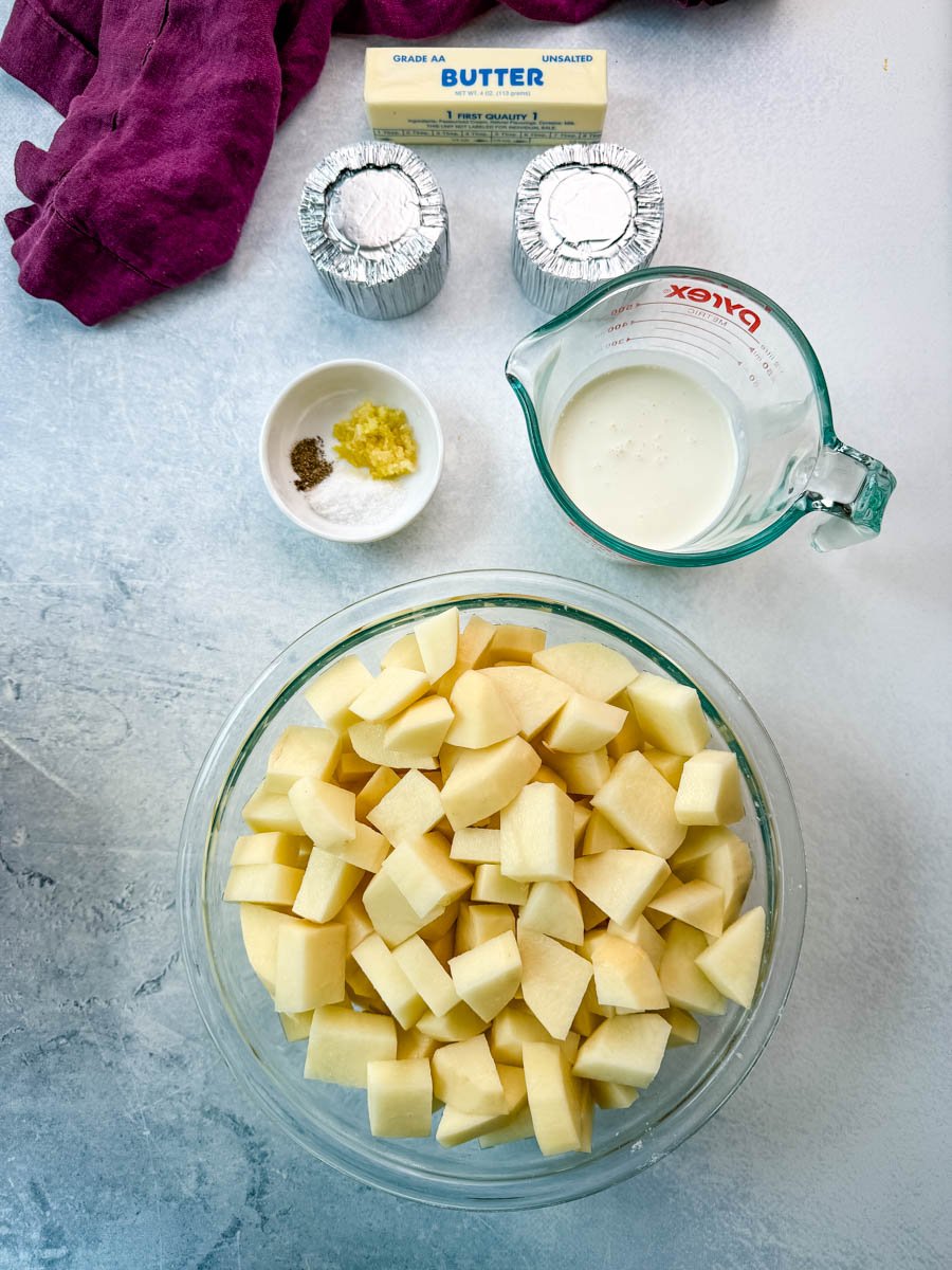 Overhead shot of peeled and cubed potatoes in a large glass bowl, surrounded by a measuring cup of milk, two foil-wrapped rounds of cheese, a stick of unsalted butter, and a small bowl with minced garlic, salt, and pepper, all arranged on a light countertop with a purple kitchen towel.