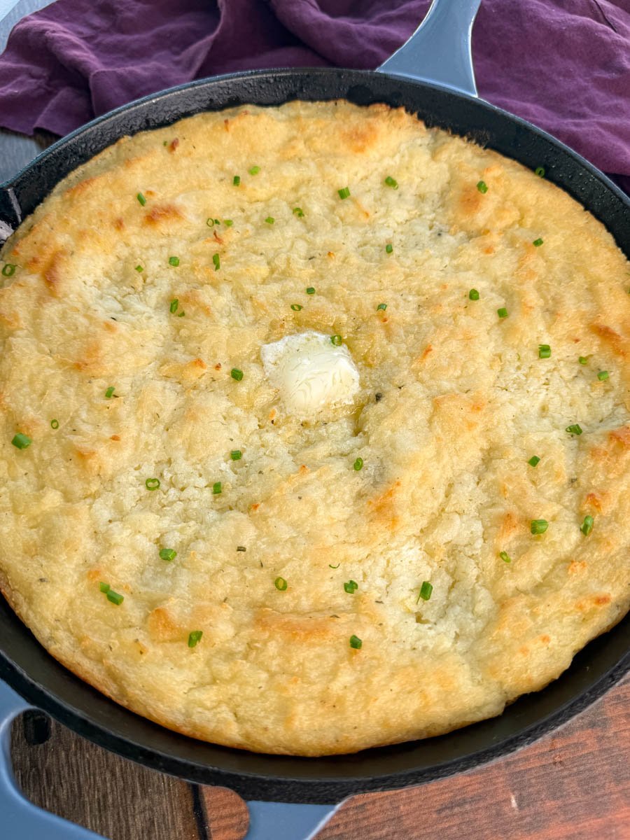 Overhead view of baked mashed potatoes in a skillet, garnished with chives and topped with a melting pat of butter in the center.