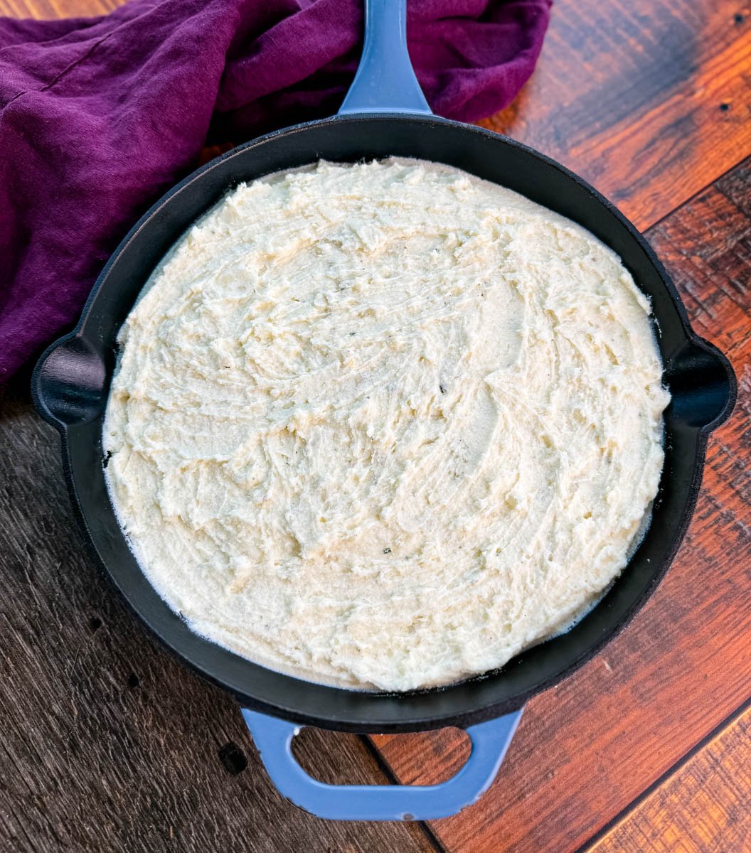Overhead view of unbaked mashed potatoes spread evenly in a cast-iron skillet, ready for the oven, with a purple cloth in the background.