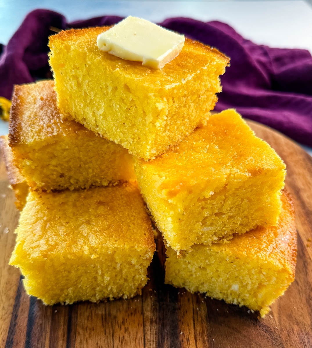A stack of golden, fluffy cornbread squares on a wooden board, topped with a pat of melting butter, with a purple cloth in the background.