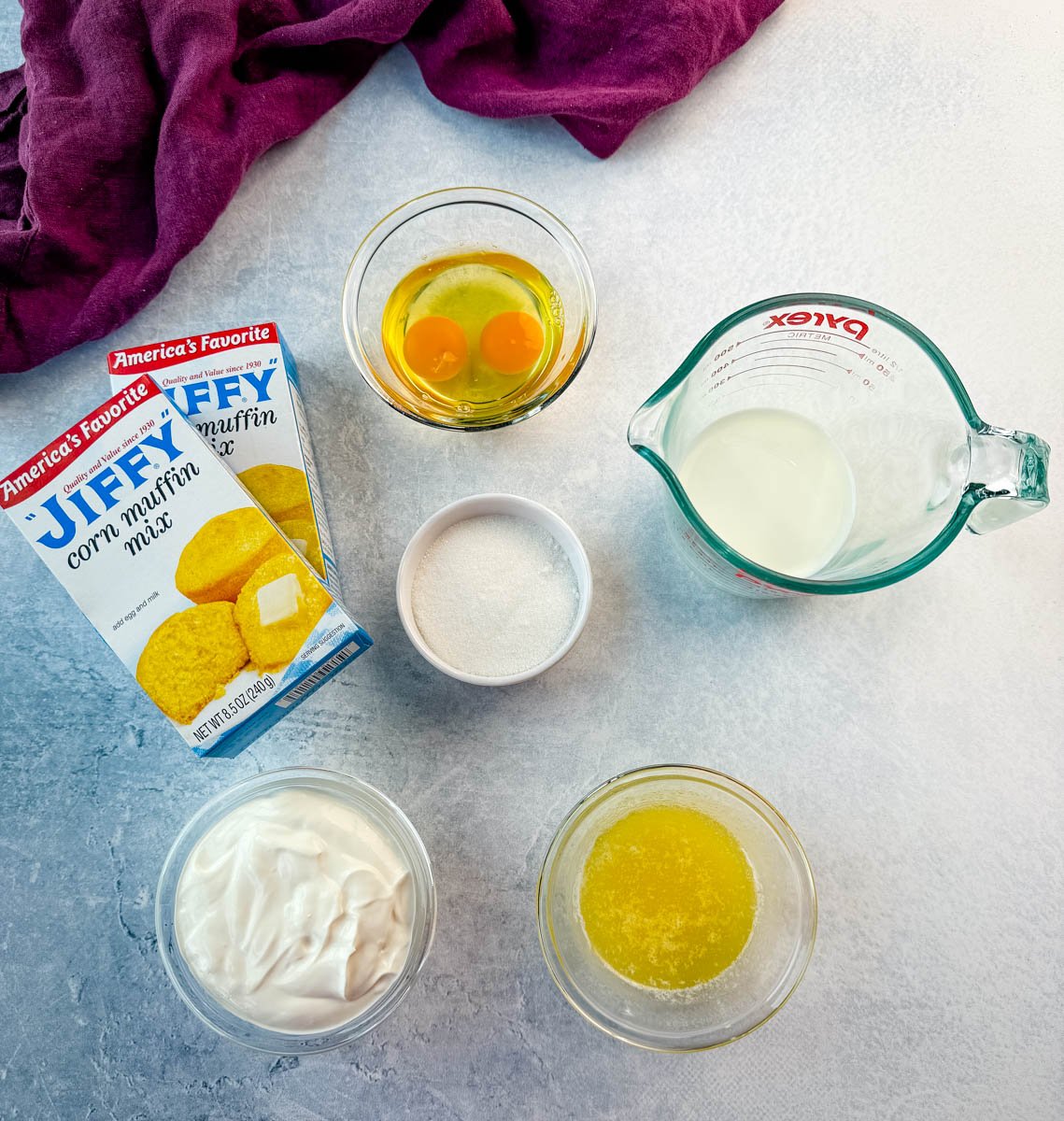 Overhead view of Jiffy cornbread ingredients on a light gray surface, including two boxes of Jiffy corn muffin mix, two cracked eggs in a glass bowl, a measuring cup of milk, a small bowl of sugar, a bowl of sour cream, and a bowl of melted butter, with a purple cloth in the background.