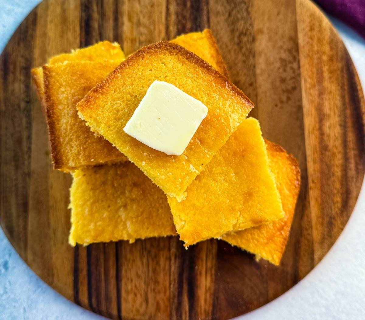 Overhead view of stacked cornbread squares on a round wooden board, the top piece topped with a square of butter.