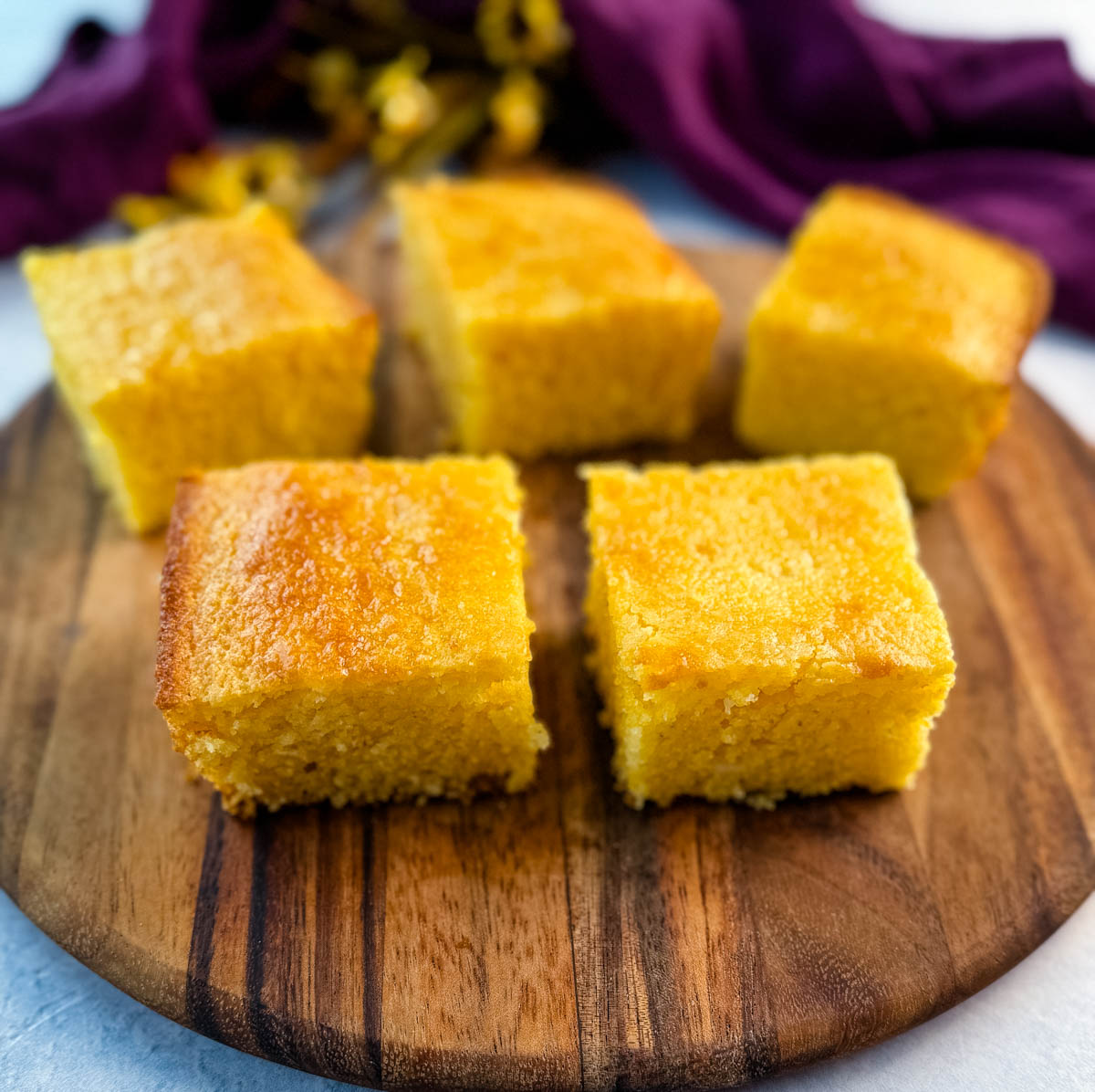 Five golden cornbread squares arranged in a circle on a round wooden board with a purple cloth and yellow flowers in the background.