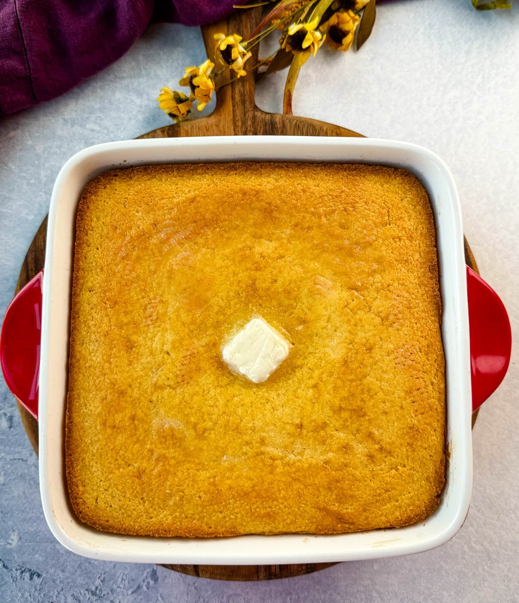 Freshly baked golden cornbread in a square white baking dish with red handles, topped with a pat of butter in the center.