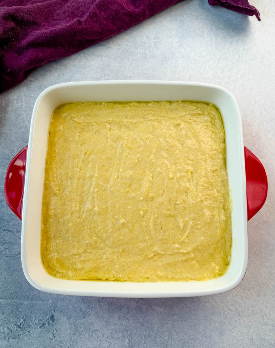 Unbaked cornbread batter spread evenly in a square white baking dish with red handles, ready for the oven, purple cloth in the background.
