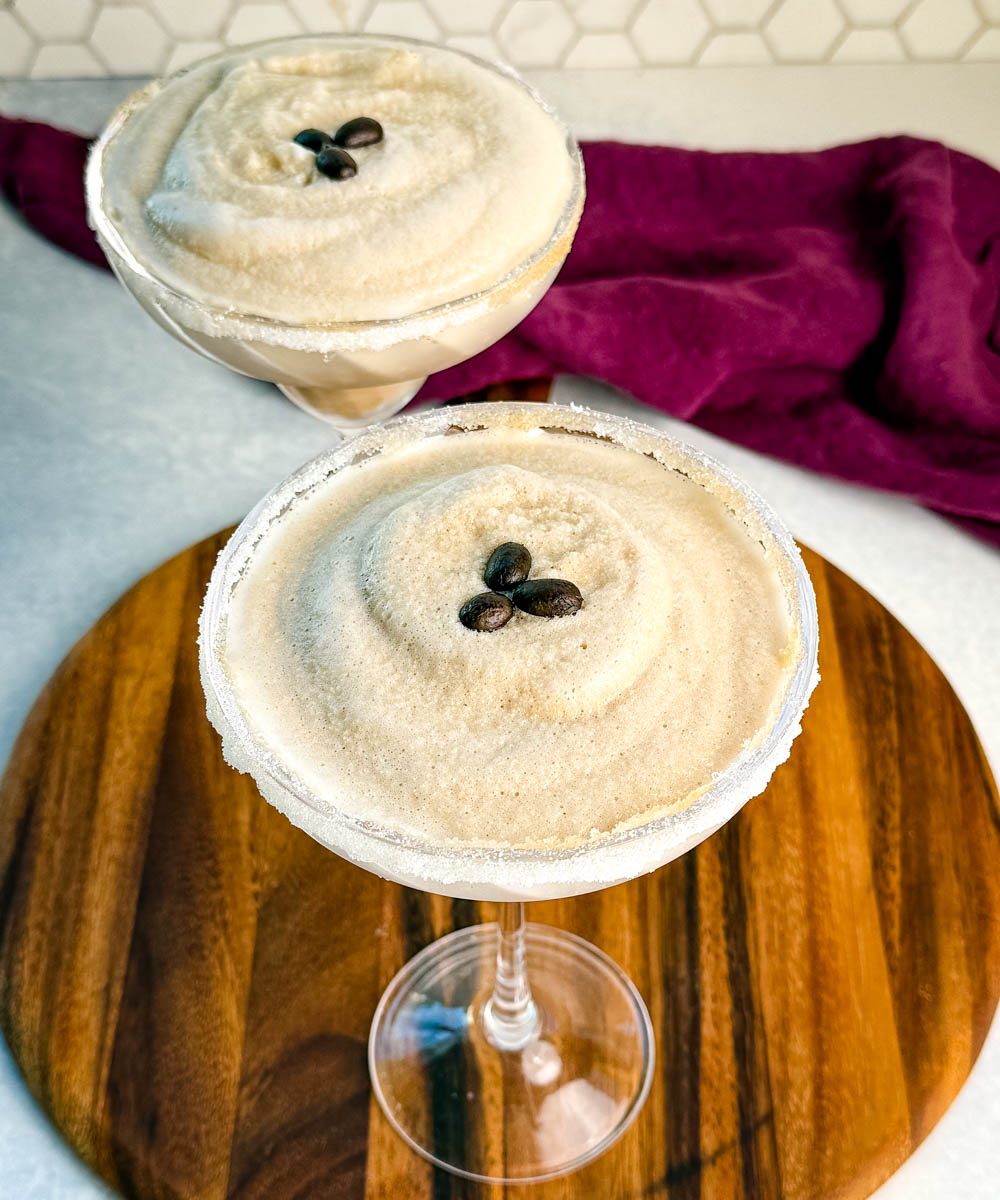Two frozen espresso martinis in sugar-rimmed glasses topped with coffee beans, served on a wooden board with a purple cloth in the background.