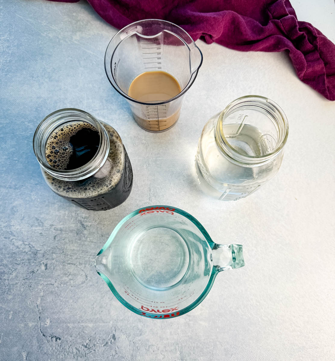 Overhead of ingredients in jars and measuring cups: vodka, coffee liqueur or Irish cream, espresso or cold brew, and water, with a purple cloth in the background. Image 9: Styled shot of two sugar-rimmed glasses filled with espresso martini slush on a wooden board, topped with coffee beans.