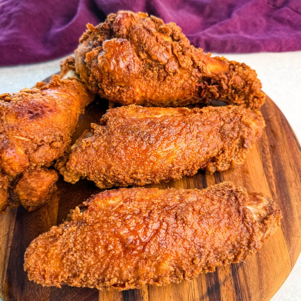 Close-up of golden brown fried turkey wings stacked on a round wooden board, with a deep purple cloth in the background.