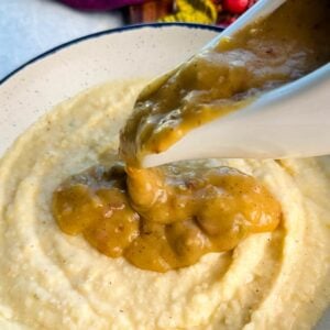 Golden turkey giblet gravy being poured generously from a white gravy boat over a bowl of creamy grits.