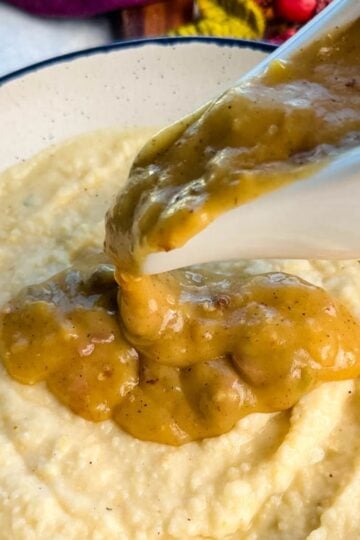 Golden turkey giblet gravy being poured generously from a white gravy boat over a bowl of creamy grits.
