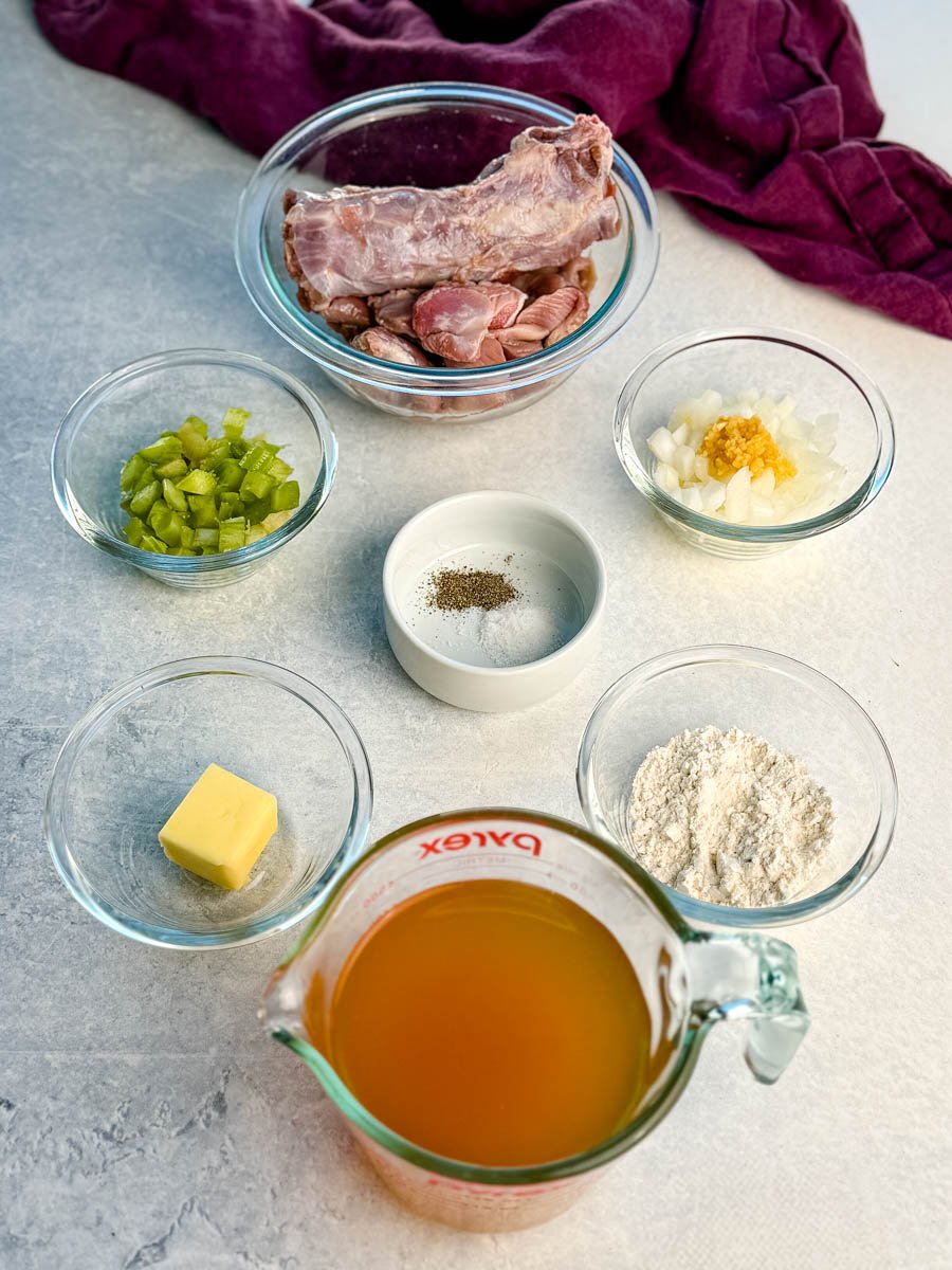 Overhead view of measured ingredients for homemade turkey giblet gravy, including raw turkey neck and giblets in a glass bowl, chopped celery, diced onions with garlic, butter, flour, chicken broth in a measuring cup, and small bowls of salt and black pepper, all arranged neatly on a light countertop with a purple cloth in the background.
