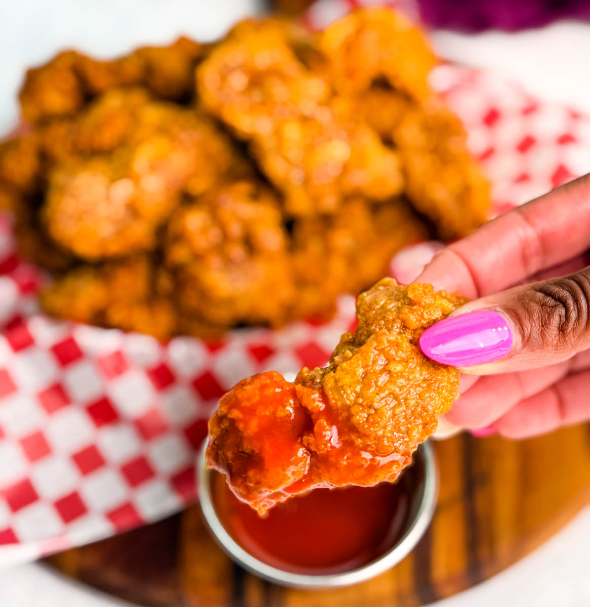 Close-up of a hand holding a crispy fried chicken gizzard dipped in bright red hot sauce, with a basket of fried gizzards blurred in the background.