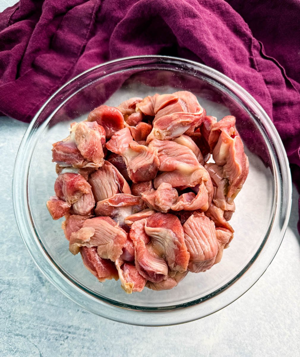 Overhead view of raw chicken gizzards in a clear glass bowl, set on a light background with a purple cloth nearby.