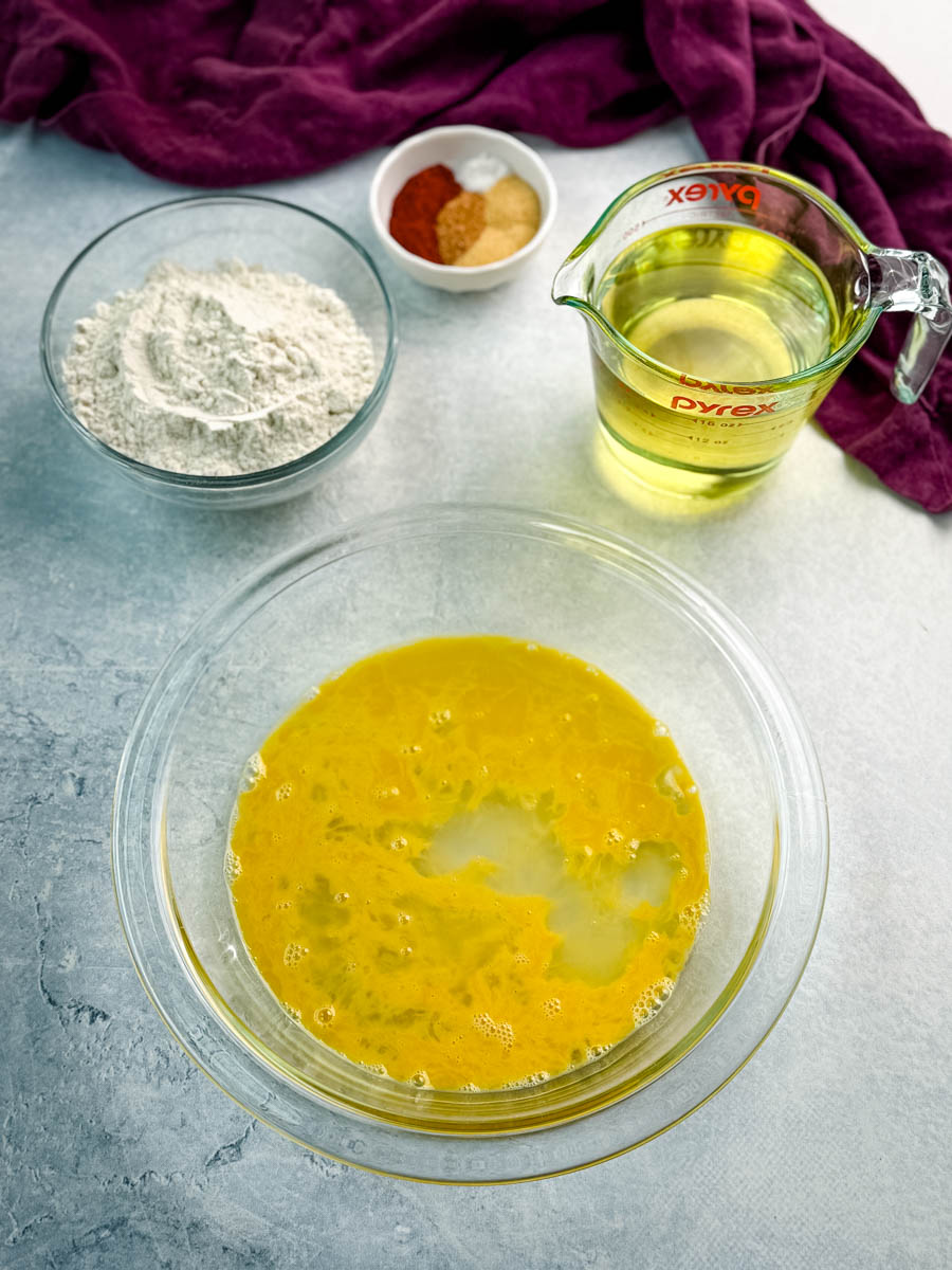 Ingredients laid out for frying gizzards, including a bowl of seasoned flour, a measuring cup of oil, a bowl of beaten egg wash, and a small dish of spices.