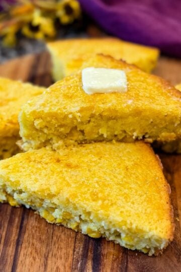 Slices of golden Jiffy corn casserole arranged on a wooden board, topped with a pat of butter, showing moist texture with visible corn kernels.