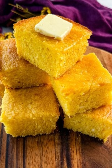 A stack of golden, fluffy cornbread squares on a wooden board, topped with a pat of melting butter, with a purple cloth in the background.