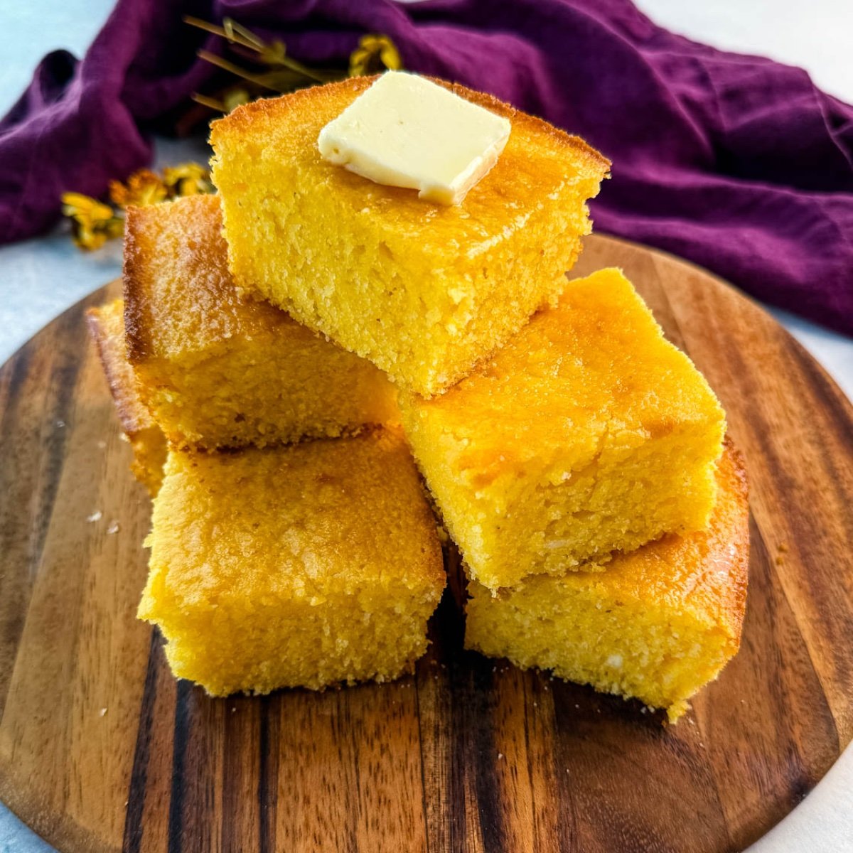 A stack of golden, fluffy cornbread squares on a wooden board, topped with a pat of melting butter, with a purple cloth in the background.