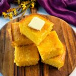 A stack of golden, fluffy cornbread squares on a wooden board, topped with a pat of melting butter, with a purple cloth in the background.
