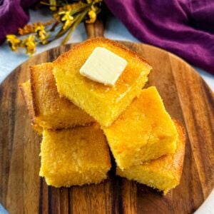 A stack of golden, fluffy cornbread squares on a wooden board, topped with a pat of melting butter, with a purple cloth in the background.