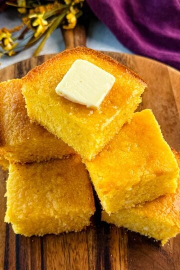A stack of golden, fluffy cornbread squares on a wooden board, topped with a pat of melting butter, with a purple cloth in the background.