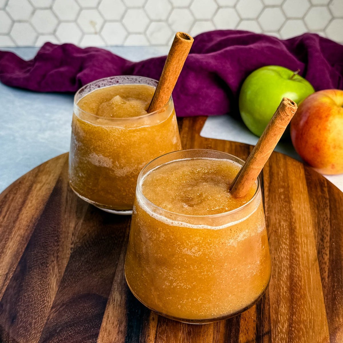 Two glasses of frozen apple cider slush served on a wooden board, each garnished with a cinnamon stick, with apples and a purple cloth in the background.