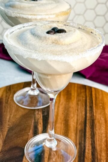 Two frozen espresso martinis in sugar-rimmed glasses topped with coffee beans, served on a wooden board with a purple cloth in the background.