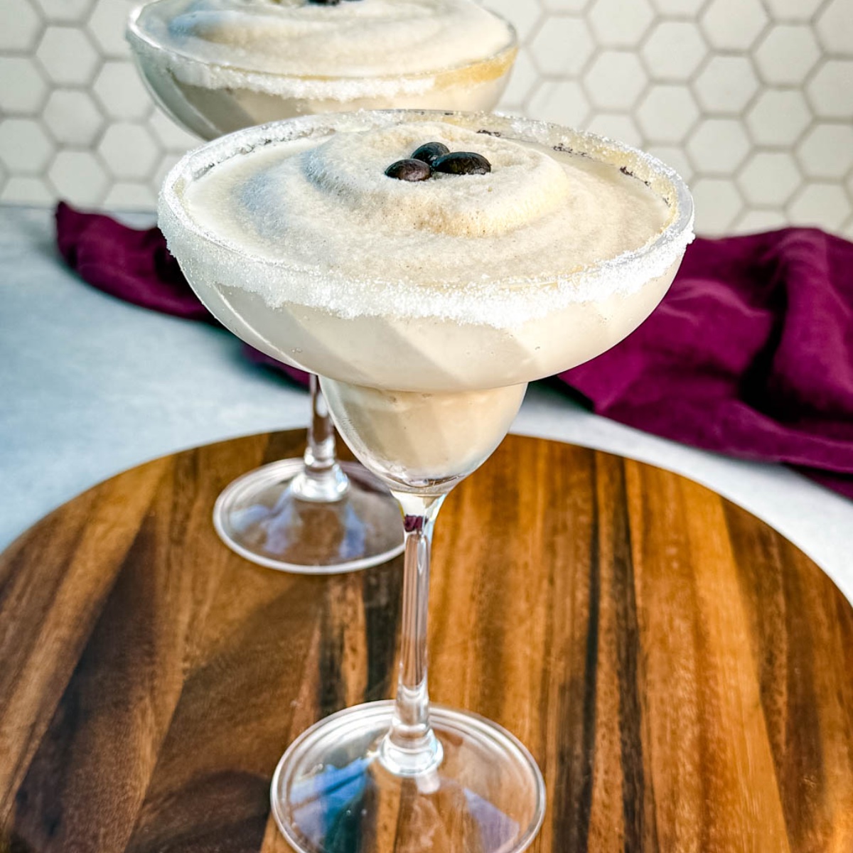 Two frozen espresso martinis in sugar-rimmed glasses topped with coffee beans, served on a wooden board with a purple cloth in the background.
