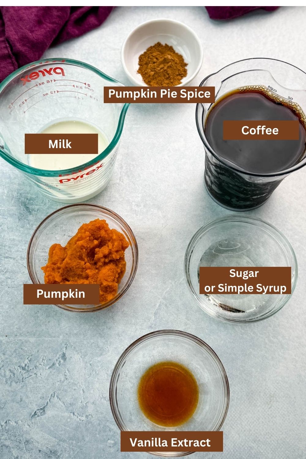 Glass bowls and measuring cups filled with milk, brewed coffee, pumpkin purée, vanilla extract, sugar or simple syrup, and pumpkin pie spice arranged on a countertop.