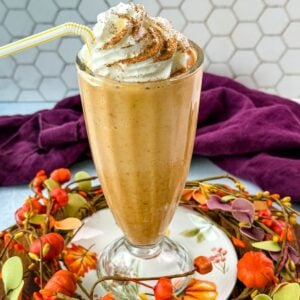 A tall glass filled with pumpkin spice milkshake topped with whipped cream and sprinkled with cinnamon, sitting on a fall-themed wooden tray decorated with small pumpkins and leaves.