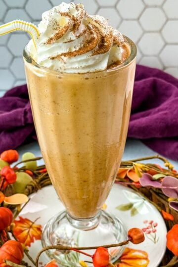 A tall glass filled with pumpkin spice milkshake topped with whipped cream and sprinkled with cinnamon, sitting on a fall-themed wooden tray decorated with small pumpkins and leaves.