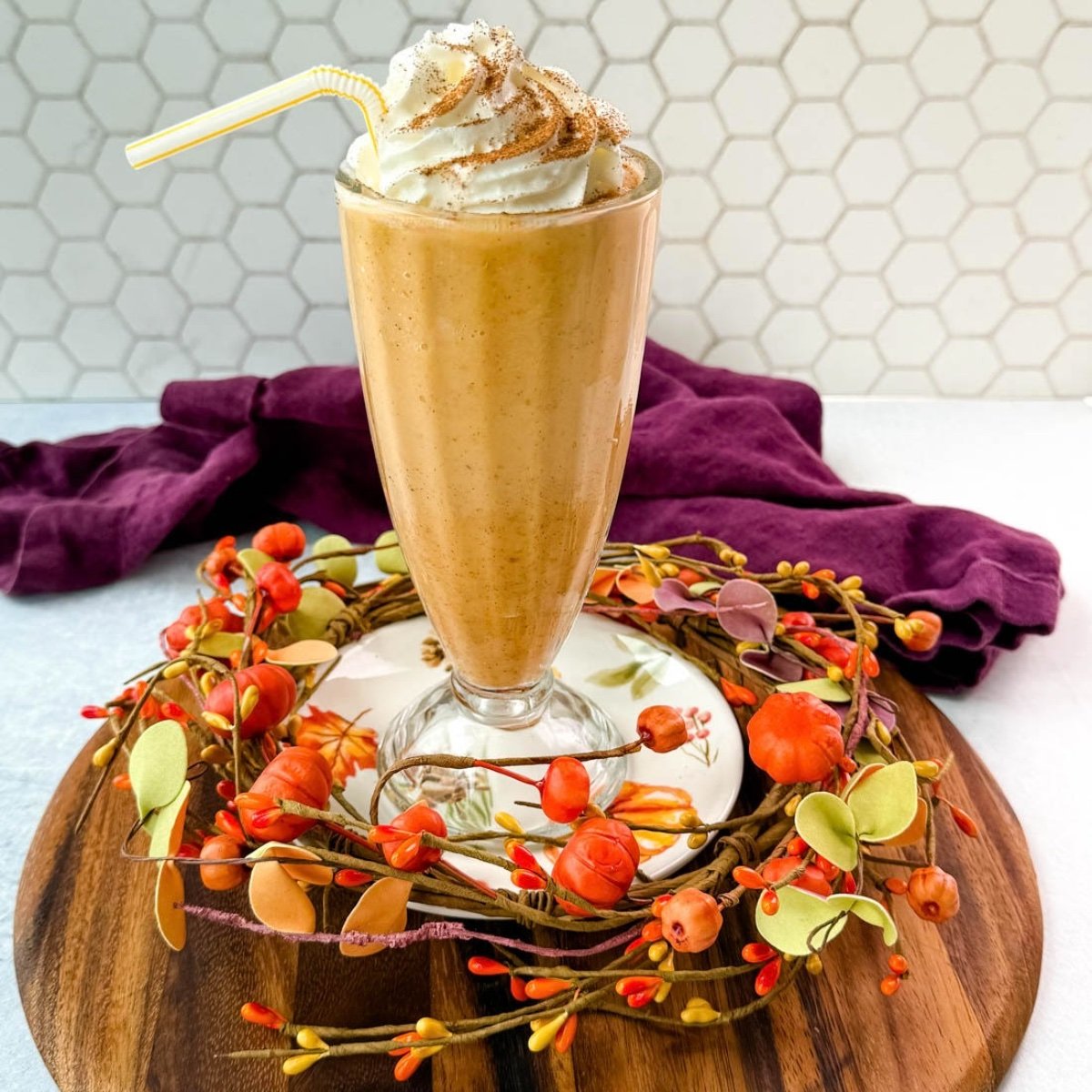 A tall glass filled with pumpkin spice milkshake topped with whipped cream and sprinkled with cinnamon, sitting on a fall-themed wooden tray decorated with small pumpkins and leaves.