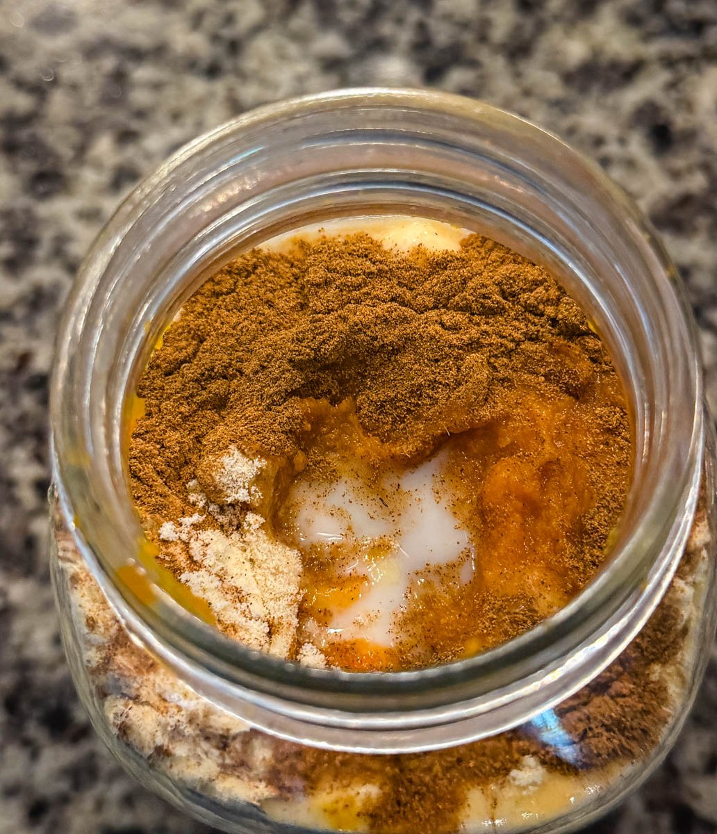 A glass jar filled with milk, pumpkin puree, protein powder, sugar, and pumpkin pie spice before blending.