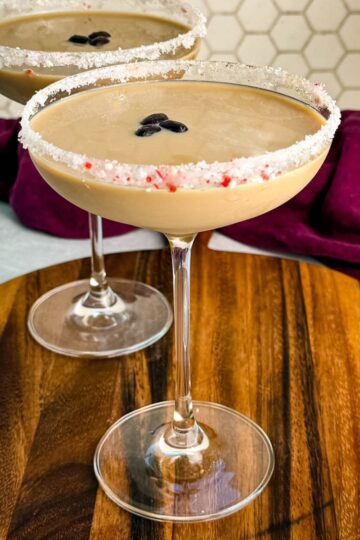 Side view of two peppermint espresso martinis in coupe glasses with crushed peppermint rims and coffee bean garnish, set on a round wooden board with a hexagon tile backsplash.