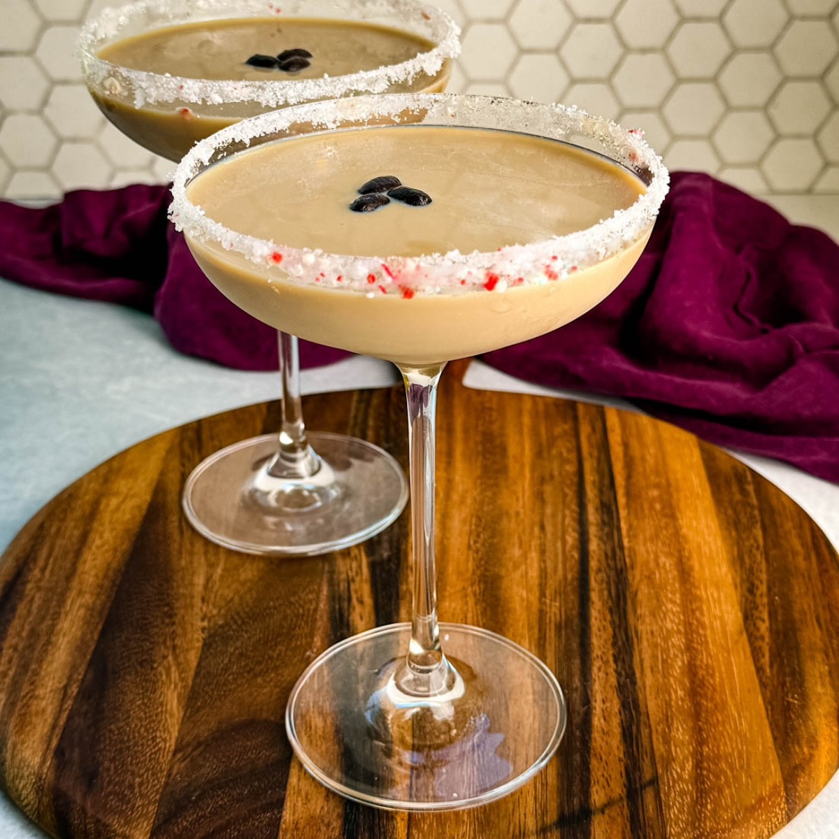 Side view of two peppermint espresso martinis in coupe glasses with crushed peppermint rims and coffee bean garnish, set on a round wooden board with a hexagon tile backsplash.
