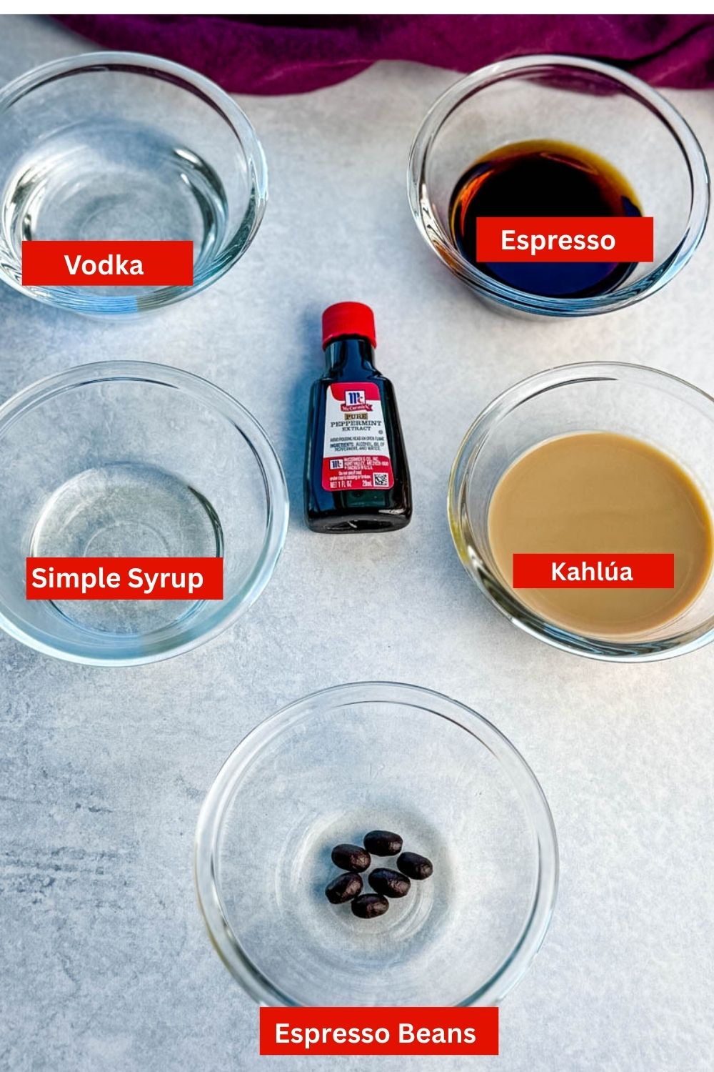 Small glass bowls with ingredients for peppermint espresso martinis including vodka, coffee liqueur, Baileys, peppermint extract, and coffee beans arranged neatly on a gray surface.