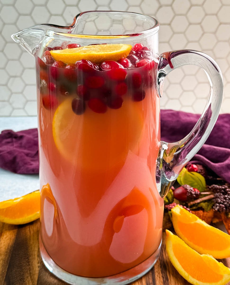 Glass pitcher filled with red holiday punch, garnished with floating cranberries and orange slices, set on a wooden board with fresh orange wedges.
