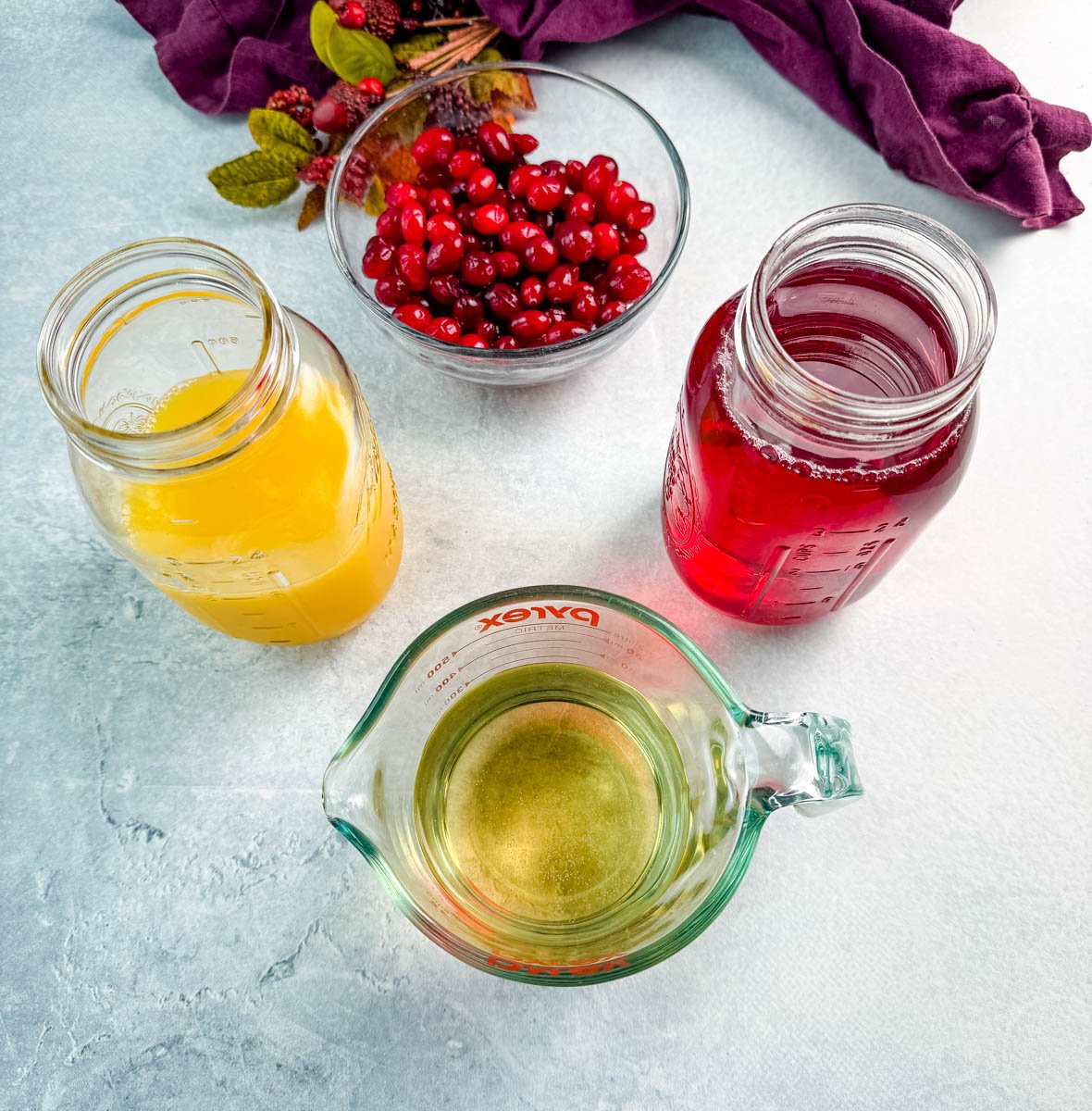 Overhead view of Christmas punch ingredients including a jar of orange juice, a jar of cranberry juice, a bowl of fresh cranberries, and a measuring cup of lemon-lime soda.