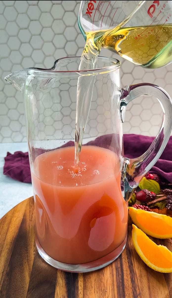Lemon-lime soda/ginger ale being poured into a pitcher already filled with punch mixture, set on a wooden board with orange wedges nearby.