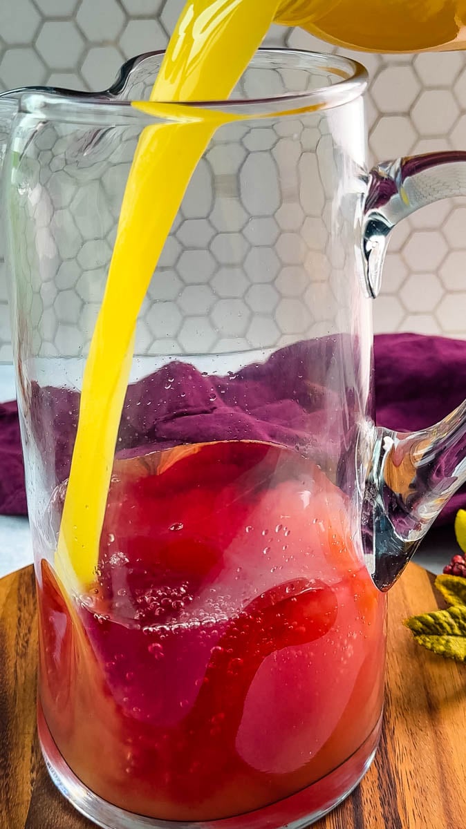 Bright orange juice being poured into a pitcher with cranberry juice, creating a colorful swirl as the liquids combine.