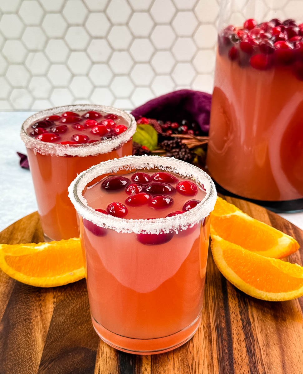 Two glasses of Christmas punch with sugared rims, each garnished with cranberries, placed in front of a large pitcher of punch.