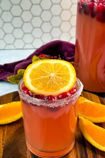 Glass of Christmas punch with sugared rim, garnished with cranberries and an orange slice, with a pitcher of punch in the background.