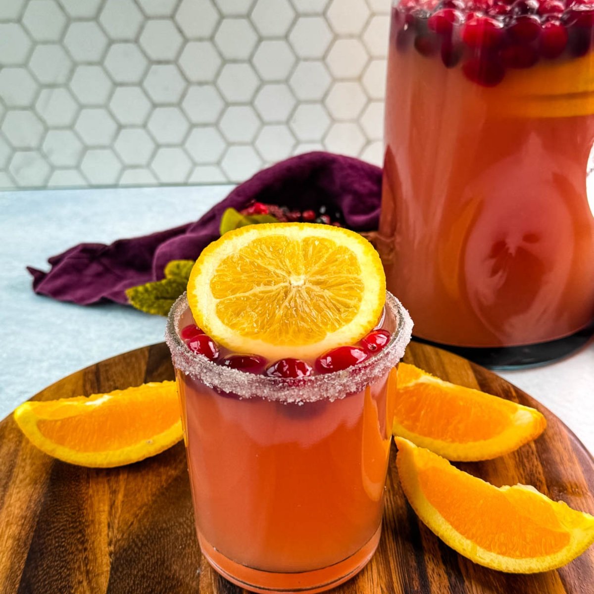 Glass of Christmas punch with sugared rim, garnished with cranberries and an orange slice, with a pitcher of punch in the background.