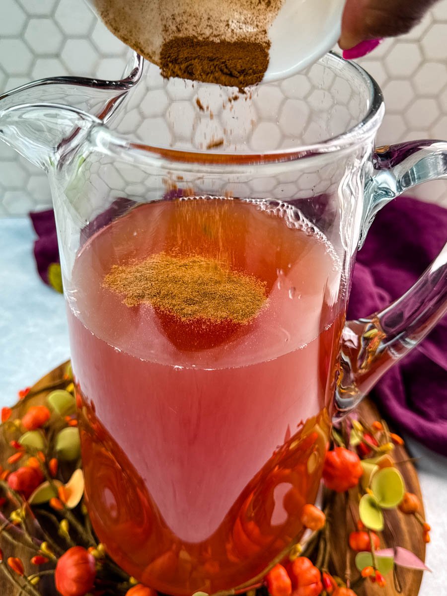 Bowl of ground cinnamon being sprinkled into a pitcher of punch, creating a warm spiced base for the holiday drink.