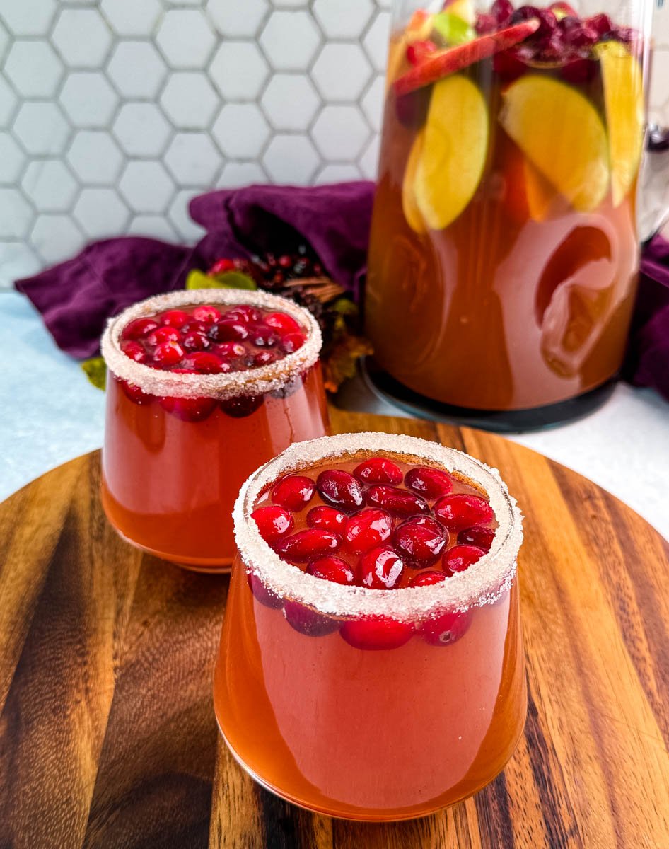 Two glasses of Thanksgiving punch with sugared rims and cranberries in front of a pitcher of punch filled with apples and cranberries.