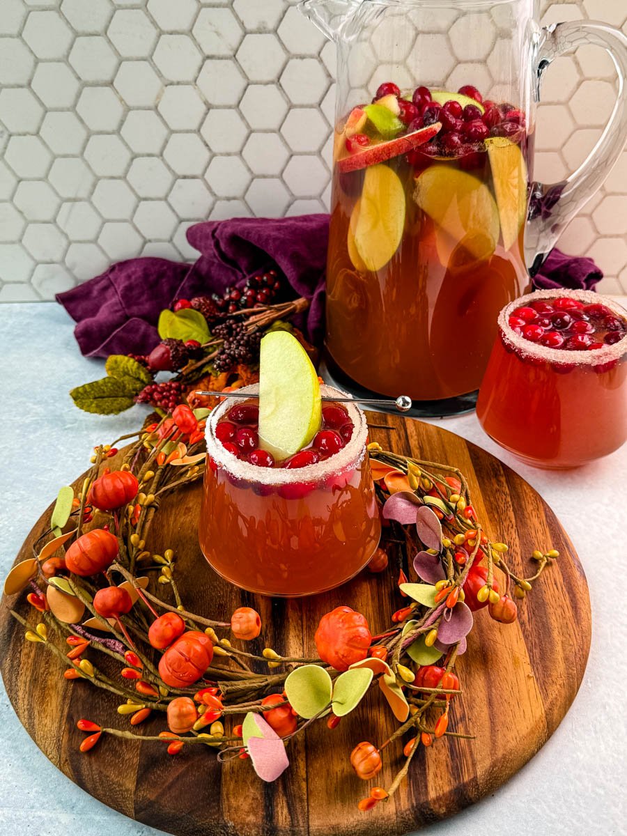 Glass of Thanksgiving punch with sugared rim, cranberries, and a green apple slice garnish in front of a pitcher filled with punch, apples, and cranberries, styled with a fall wreath on a wooden board.
