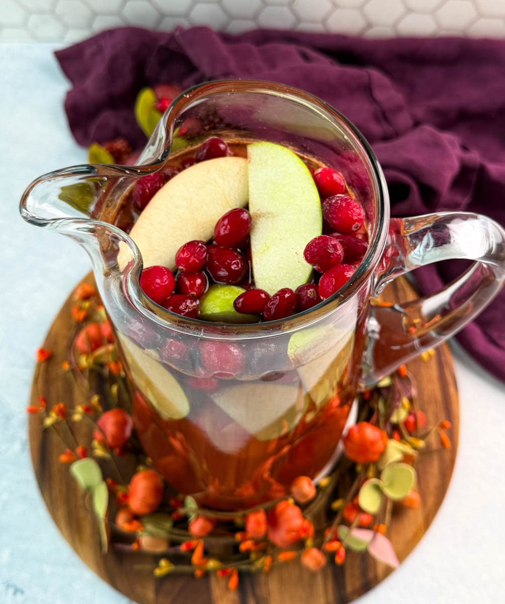 Overhead view of a pitcher of Thanksgiving punch filled with green apple slices and fresh cranberries, surrounded by autumn décor.