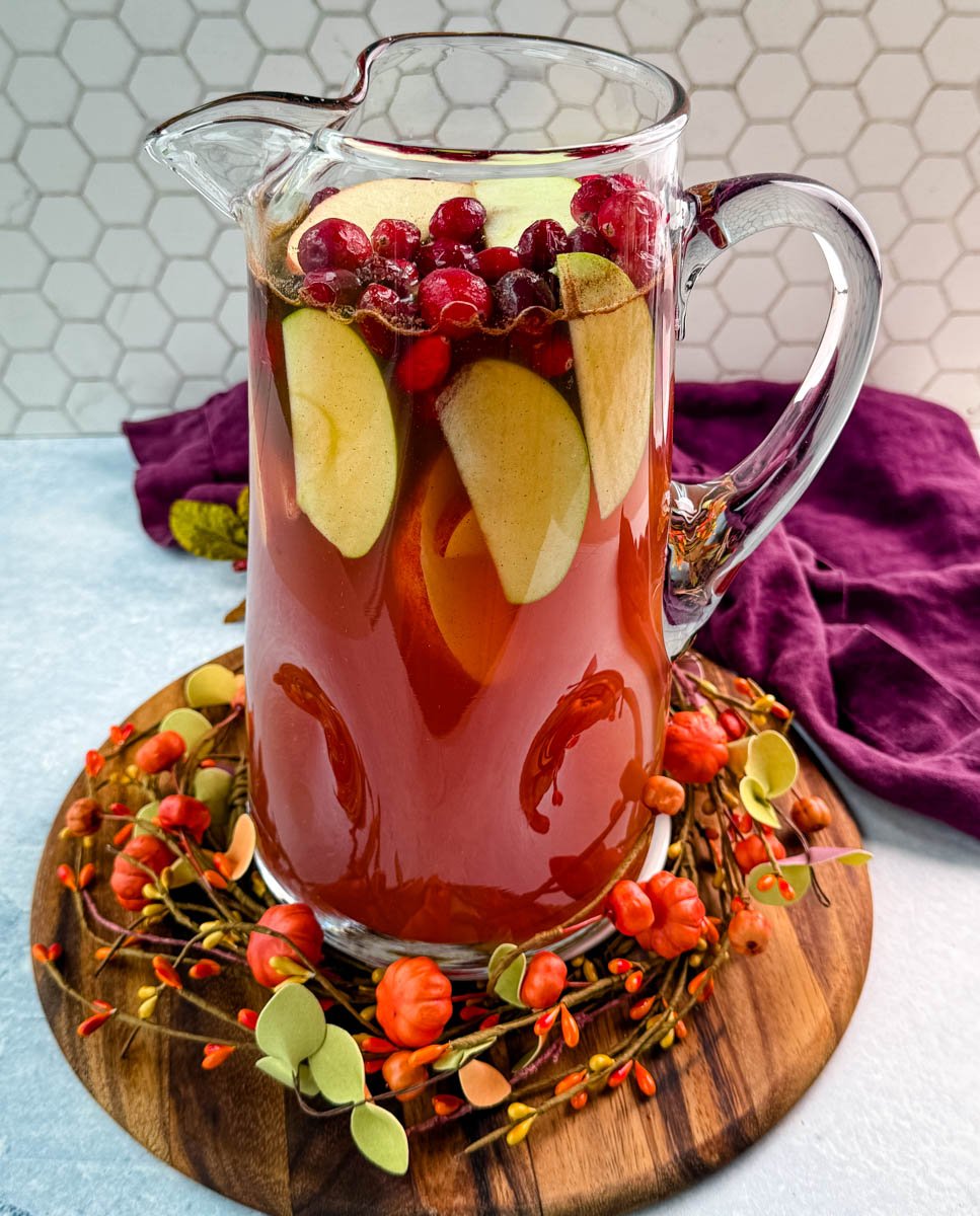 Tall pitcher of Thanksgiving punch filled with apple slices and cranberries, sitting on a fall wreath centerpiece.